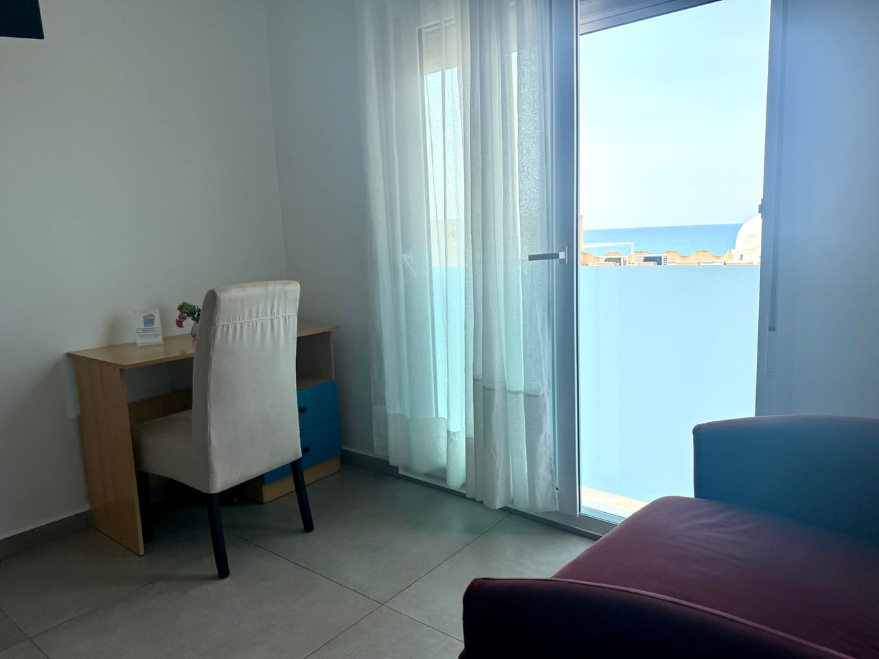 Habitación luminosa con escritorio y sillón, la puerta de la terraza abierta al mar Mediterráneo